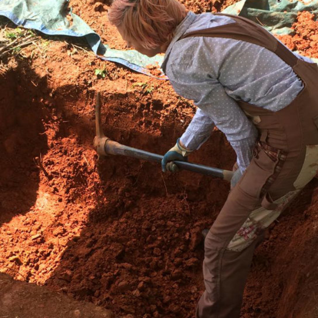 Cassie standing in a grave of red clay, using a pickaxe.