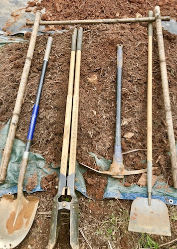 A shovel, post hole digger, mattock, and edger lying on the ground.