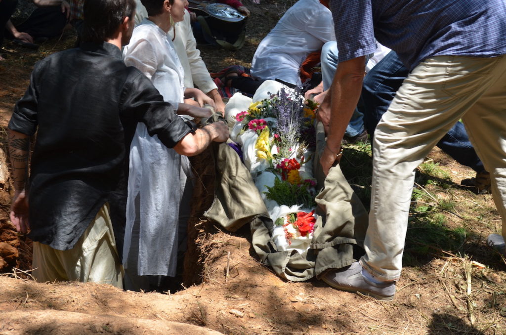 two people standing in a grave with a few people standing above, getting ready to lower a shrouded body