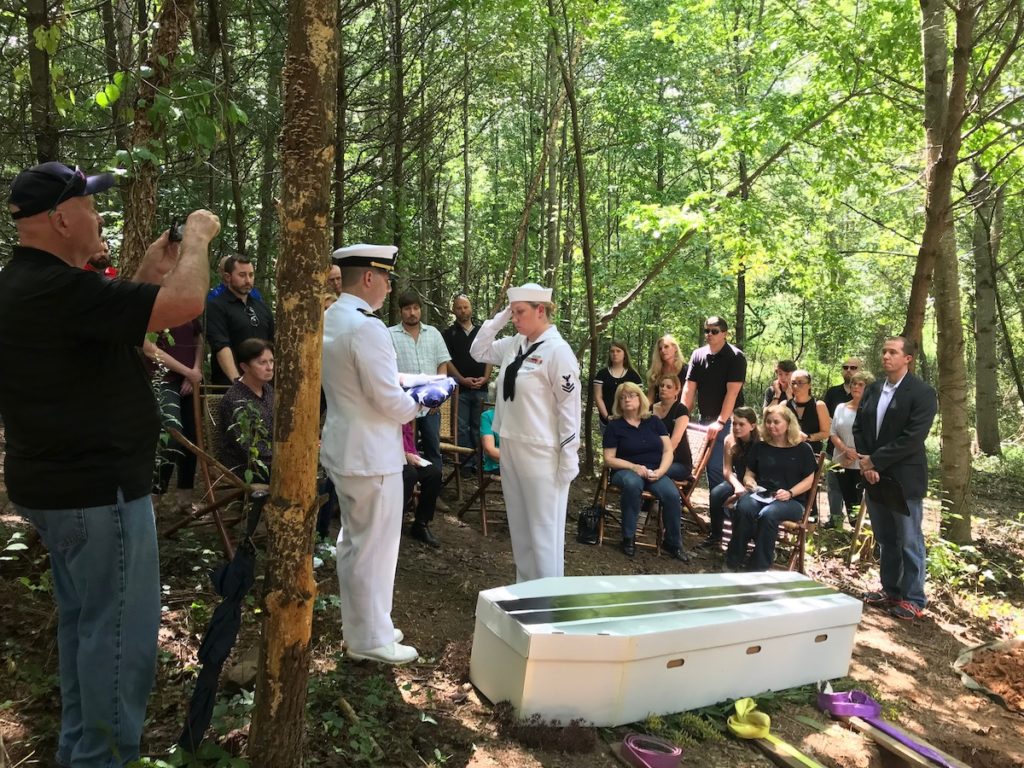 folding of the flag next to a cardboard coffin with people in the background