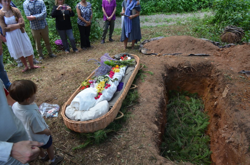 a shrouded person lying beside an open grave with pine bows in the bottom