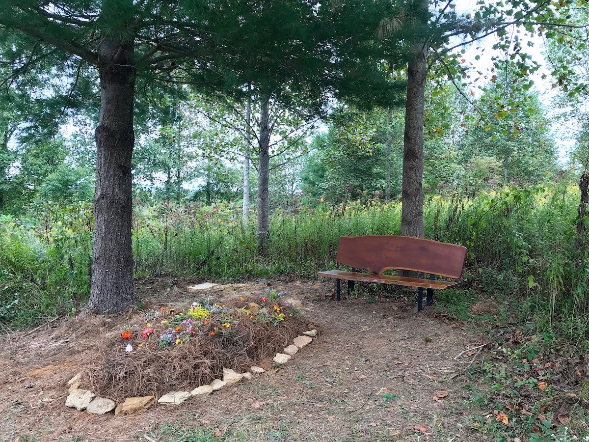 grave with flowers on top beside a bench and pine tree